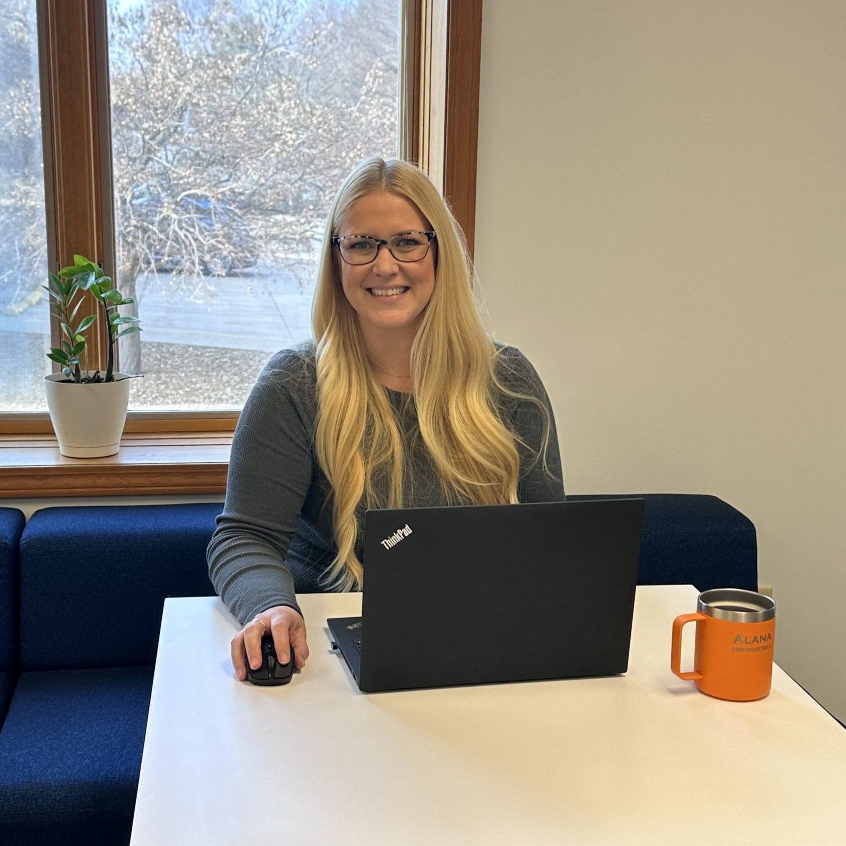 A woman smiles over a laptop at a workspace by a window