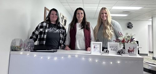 Three smiling women stand behind a desk in an office, which is decorated with twinkle lights.